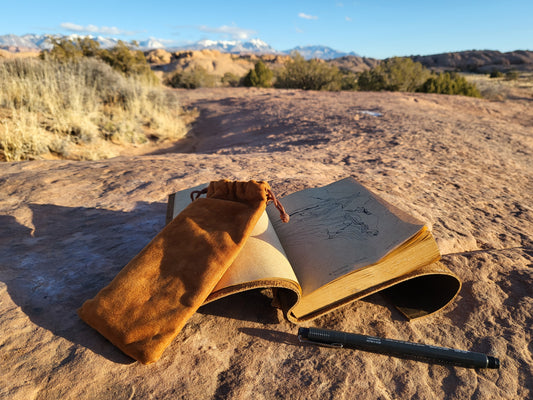 Open sketchbook with a drawing, brown pouch, and pen on sunlit rock in a desert landscape. Mountains and clear sky in the background.